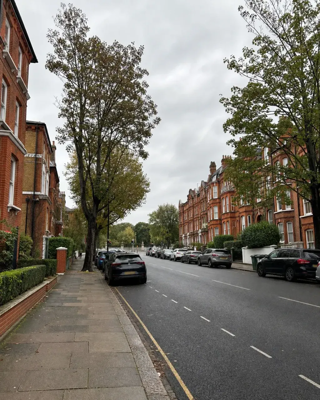 A formal avenue near Battersea Park lined with tall red brick Victorian mansion blocks, with very large mature trees and dense parkland greenery along the far side of the road